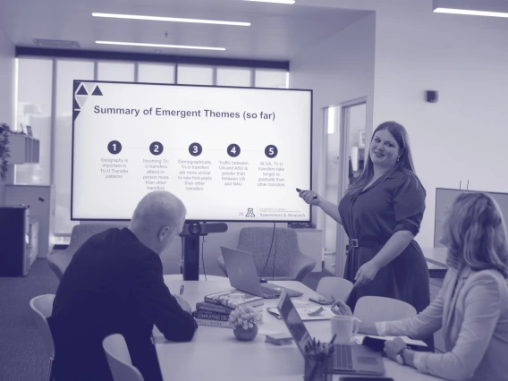three people gathered around a table with one woman presenting in front of a screen that says "key takeaways so far"